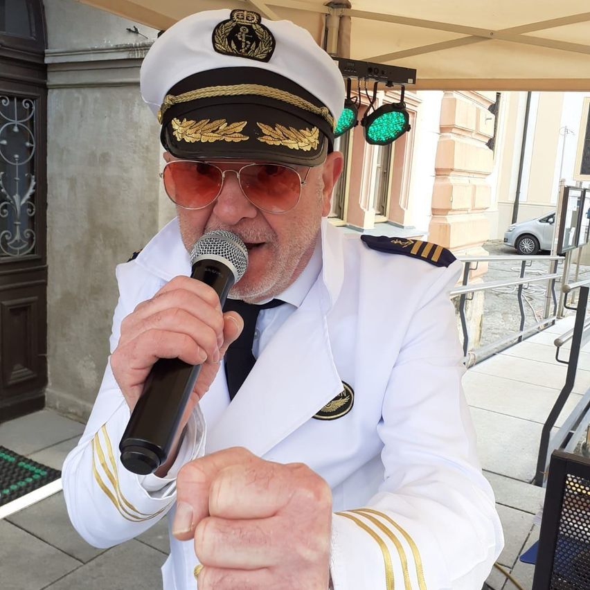 Ein Mann in einer weißen Marineuniform mit einer Mütze hält ein Mikrofon und scheint zu singen. Er steht unter einem Zelt vor einem Gebäude.