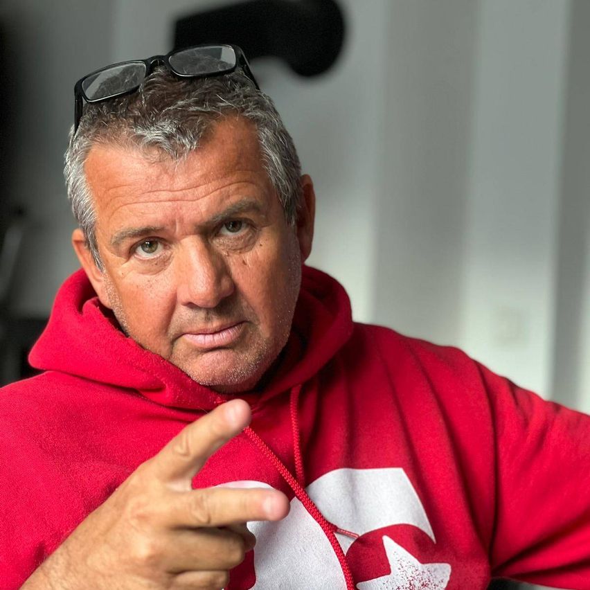 Ein Mann mit grauem Haar trägt eine rote Kapuzenjacke mit einem weißen Sternlogo auf der linken Brust. Er zeigt mit der rechten Hand auf etwas. Er trägt eine Brille auf dem Kopf.