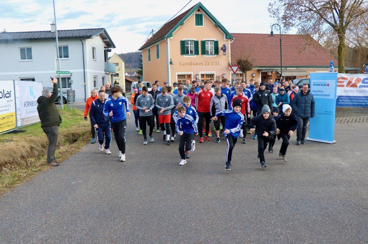 Eine Gruppe von Menschen in Sportkleidung läuft auf einer Straße, mit Häusern und einem Gebäude namens Gasthaus Zett im Hintergrund.