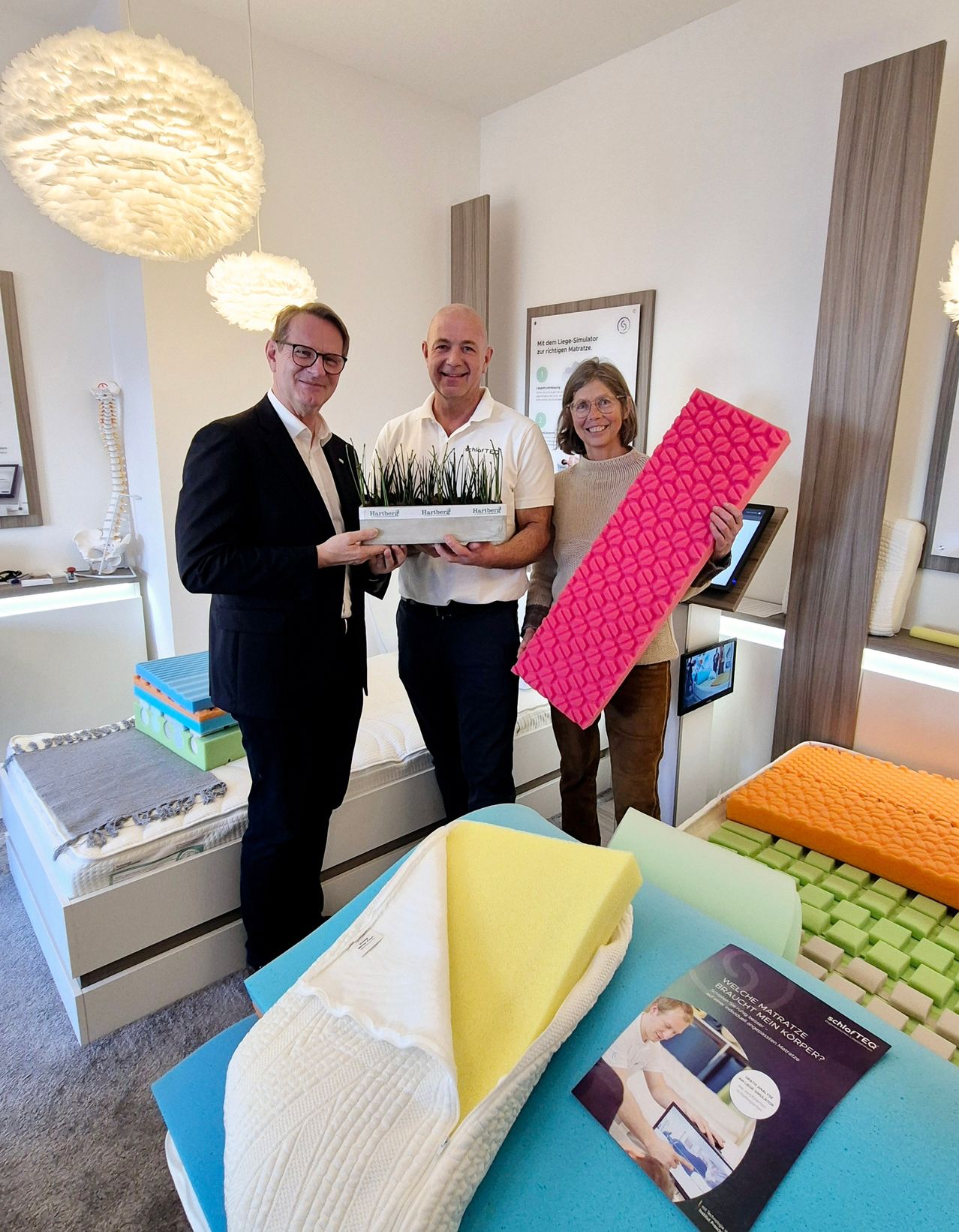Three people stand in a room with mattresses and foam products. A man in a suit holds a potted plant and another man in a polo shirt holds a pink foam mattress. A woman holds a pink foam mattress.