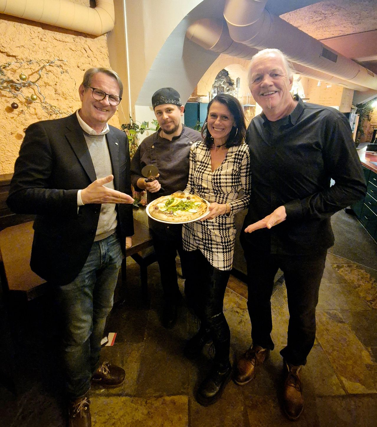 Four people are posing for a picture in a restaurant. A man is holding a pizza plate and a pizza cutter. Another man is smiling. The woman is holding the pizza plate.