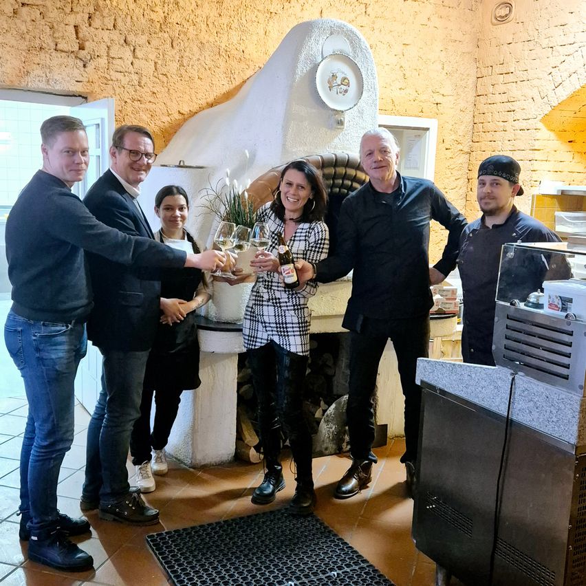 Five people are standing in a room, smiling and holding bottles and wine glasses. A woman in the middle holds a bottle and a wine glass. Behind them is a fireplace with a decorative plate. A man on the right is wearing a hat.