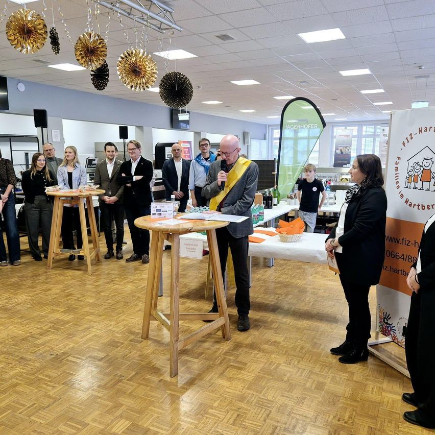 A group of people are gathered in a room with wooden floors. A man in glasses stands at a table, speaking. Two women stand in front of a banner with the website address www.fizz-hartberg.at.