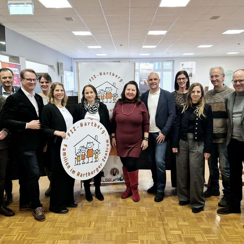 A group of people, including adults and children, stand in a room holding a circular sign. The sign features a family house logo and text. They are dressed formally and smiling for a photograph.