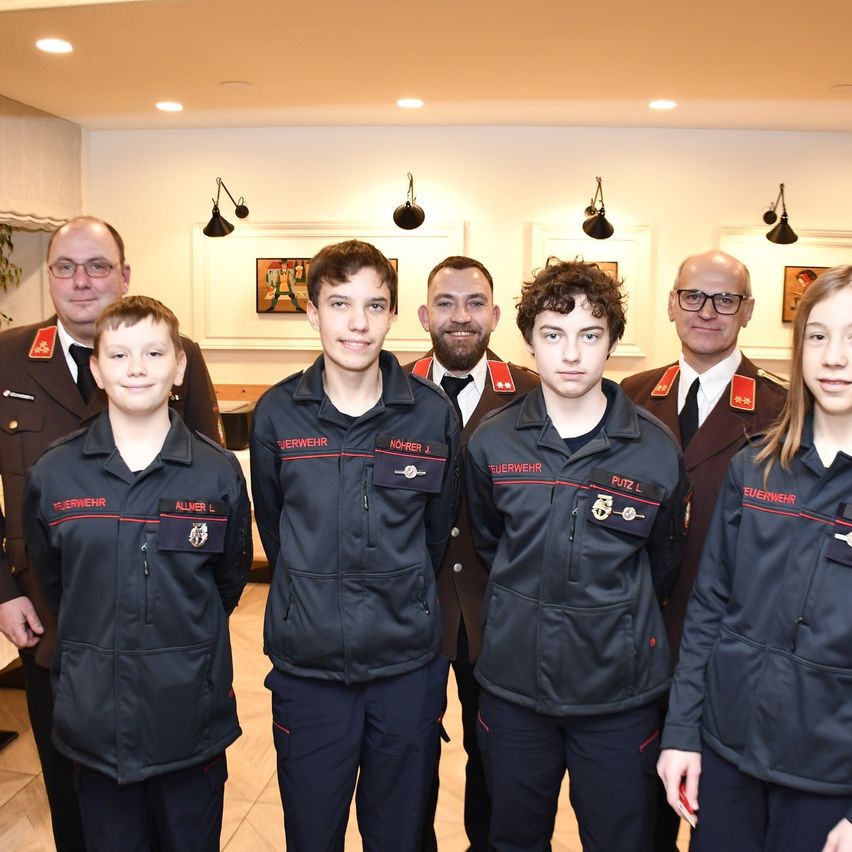 Six individuals dressed in matching jackets with red trim pose for a photo. Four of them are young boys, while the other two are older men. They are all smiling and standing together in a room.