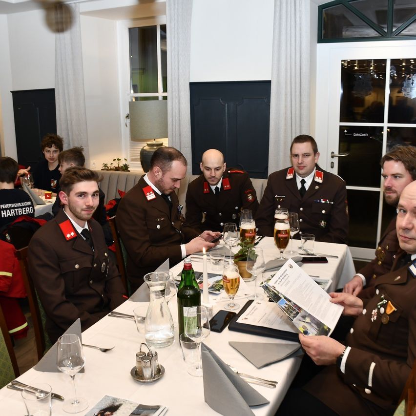 Military personnel in uniform are seated around a dining table, some are engaged in conversation while others are looking at papers on the table. There are glasses, a candle, and a green bottle on the table.
