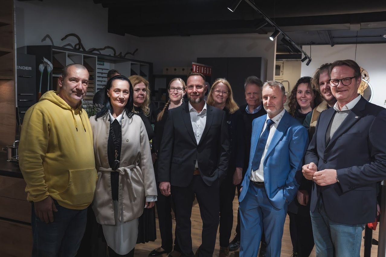 A group of people standing together in a room. One man is in a black suit, another in a blue suit. A woman in a beige coat smiles. There is a red sign on the wall.