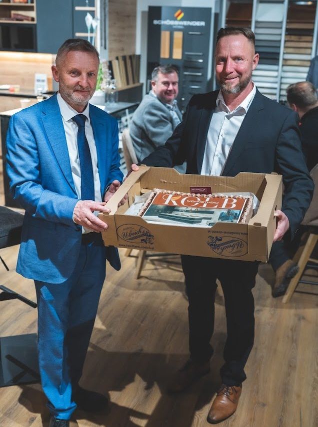 Two men hold a box with a cake inside. The man on the left wears a blue suit and tie. The man on the right wears a black suit. The cake has the word ROB on it. Behind them are two other men, one seated and one standing.