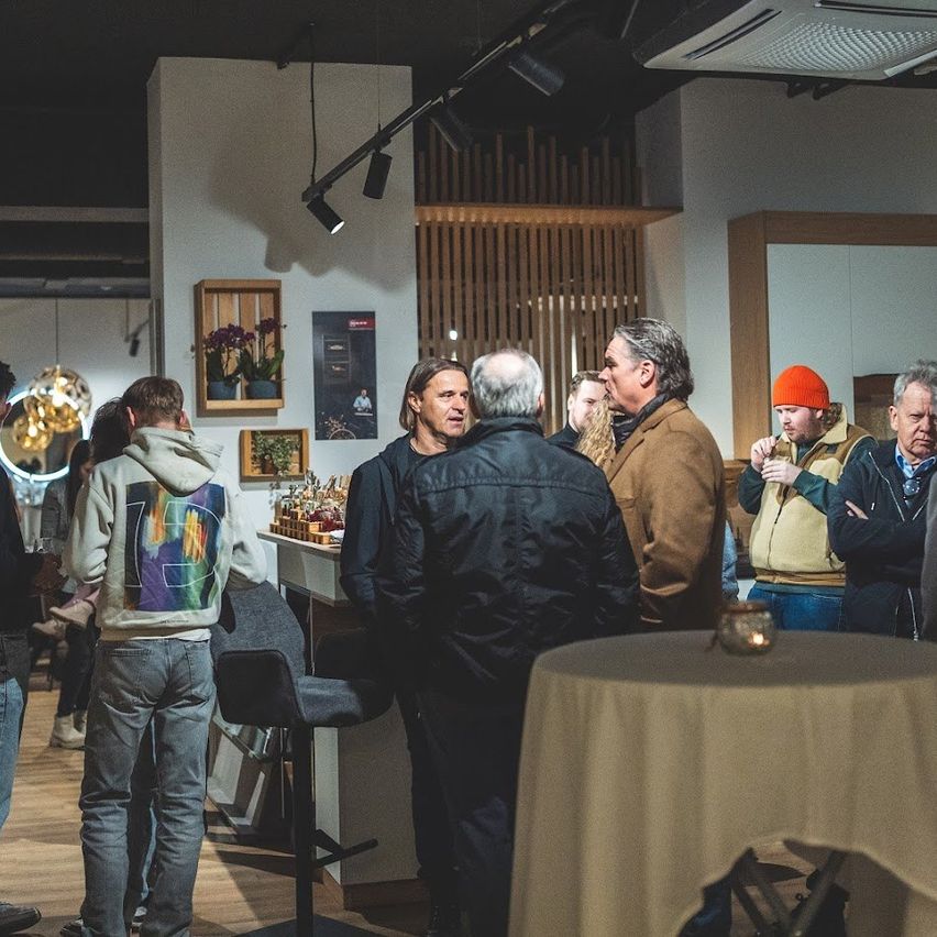 Several adults are gathered in a room, possibly a bar, with tables and chairs. One man is holding a glass.