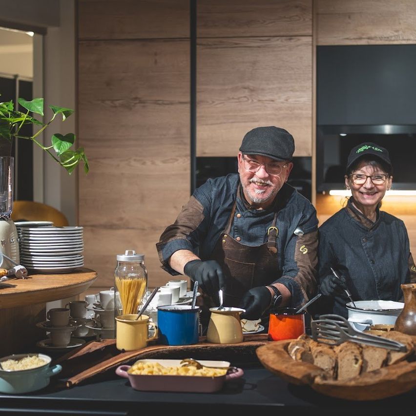 Two chefs stand behind a buffet table filled with various dishes. One is wearing a hat and apron, and the other is wearing a cap. The table has plates, bowls, and cups. A plant and plates are on a wooden table to the left.