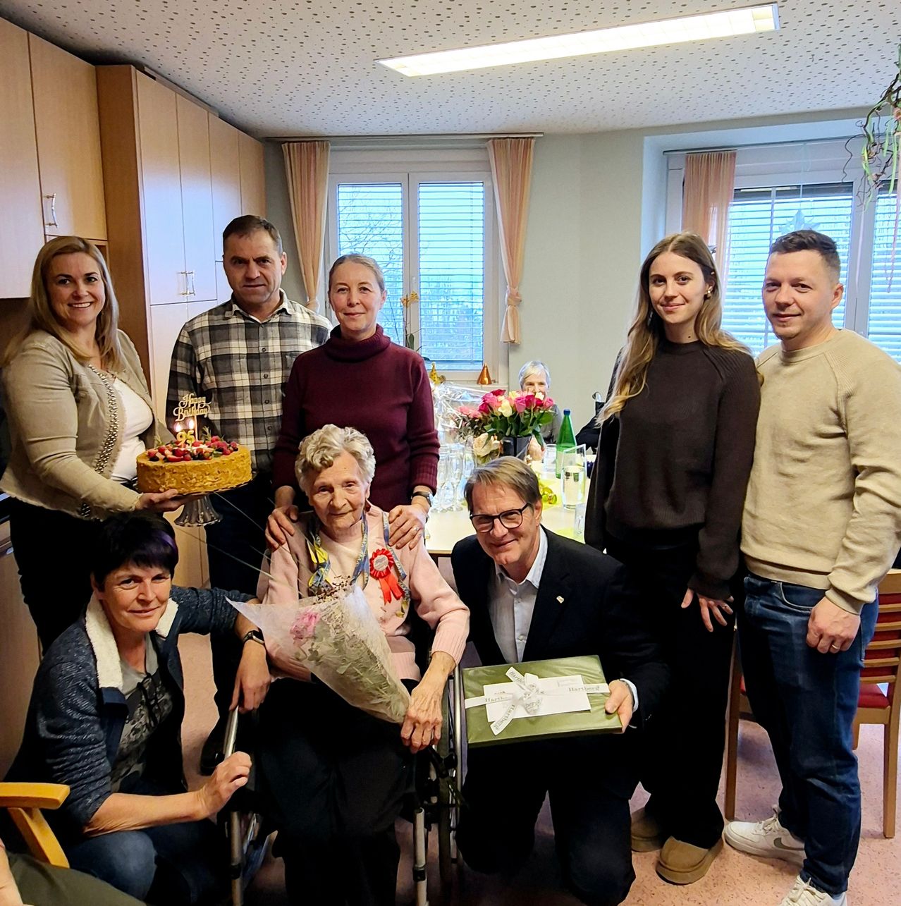 A group of people stand and sit in a room. An elderly woman in a wheelchair holds a bouquet of flowers. A man kneels and holds a gift box. A cake is held by a woman. Behind them are cabinets, a window, and a plant.