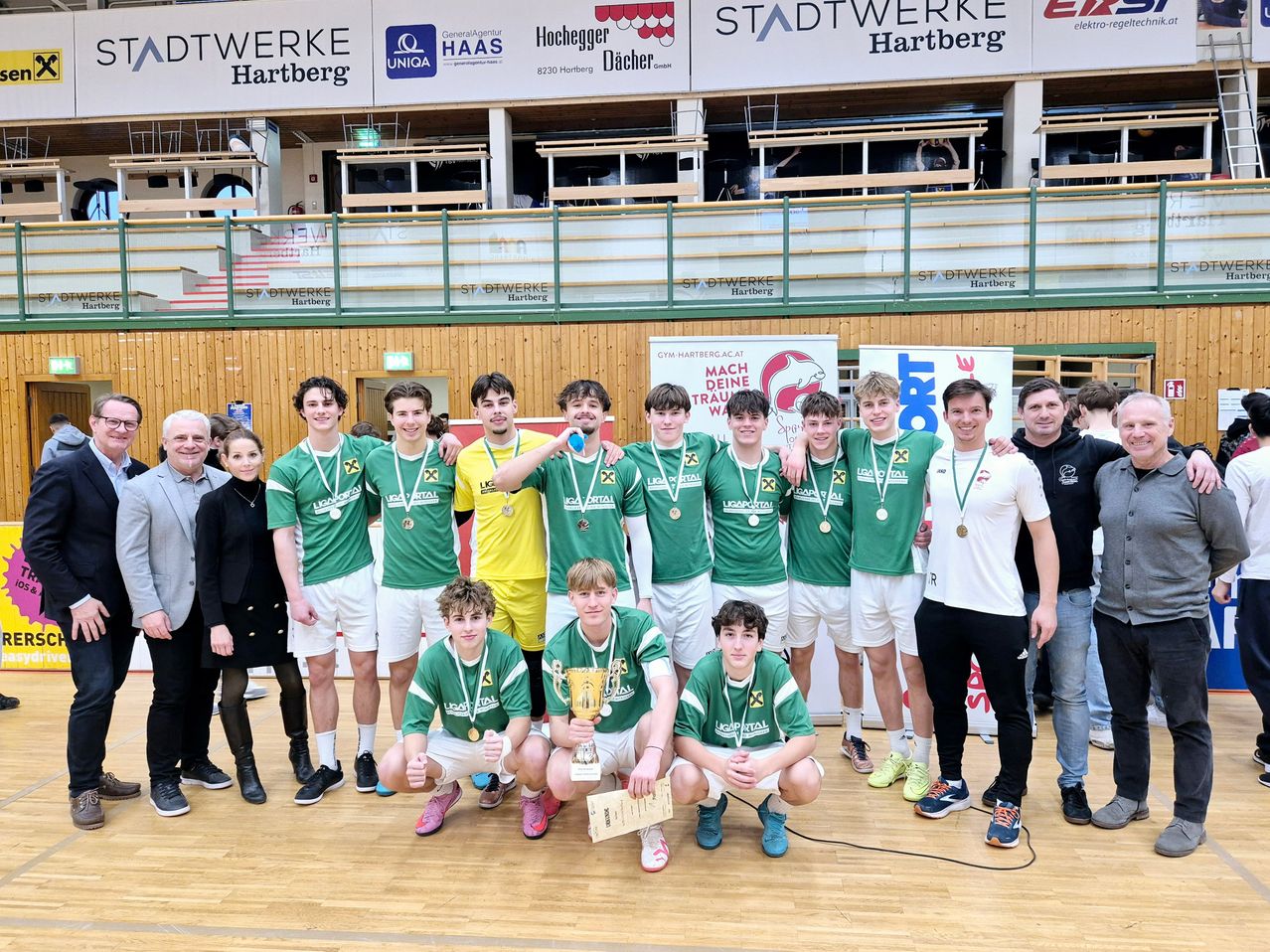 Eine Gruppe junger Männer in grünen Trikots und Medaillen posiert für ein Foto mit einer Trophäe. Dahinter stehen einige Erwachsene, einer hält eine Urkunde. Sie stehen auf einem Holzboden mit Bannern an der Wand.