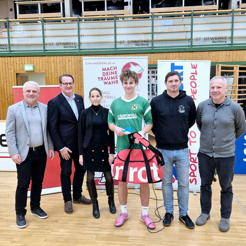 Eine Gruppe von Menschen steht in einer Turnhalle zusammen. Ein Junge hält einen roten Beutel und einen Volleyball in den Händen. Hinter ihnen sind Banner an der Wand befestigt.
