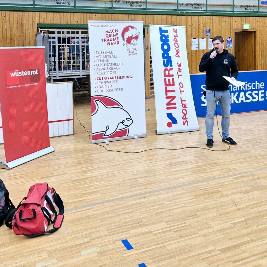 Ein Mann spricht in ein Mikrofon vor Bannern, darunter eines von 'Intersport'. In der Nähe liegt eine rote Sporttasche auf einem Holzboden.