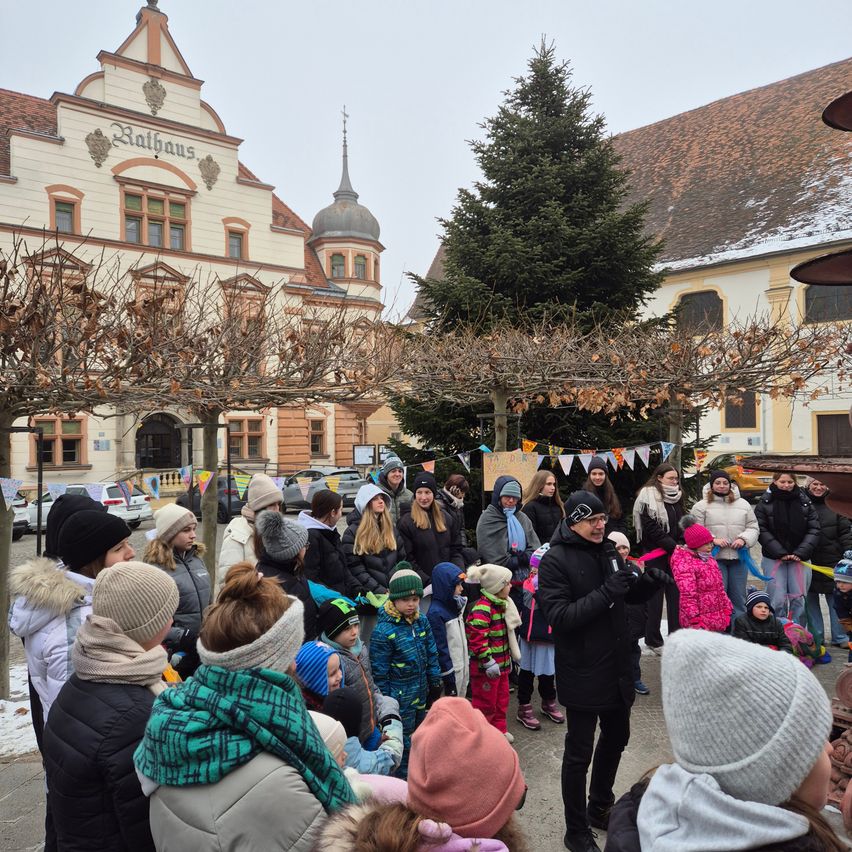 Eine große Gruppe von Menschen, darunter Erwachsene und Kinder, versammelt sich vor einem Gebäude. Sie sind warm gekleidet und einige tragen Mützen. Das Gebäude hat ein Schild mit der Aufschrift 'Rathaus'.