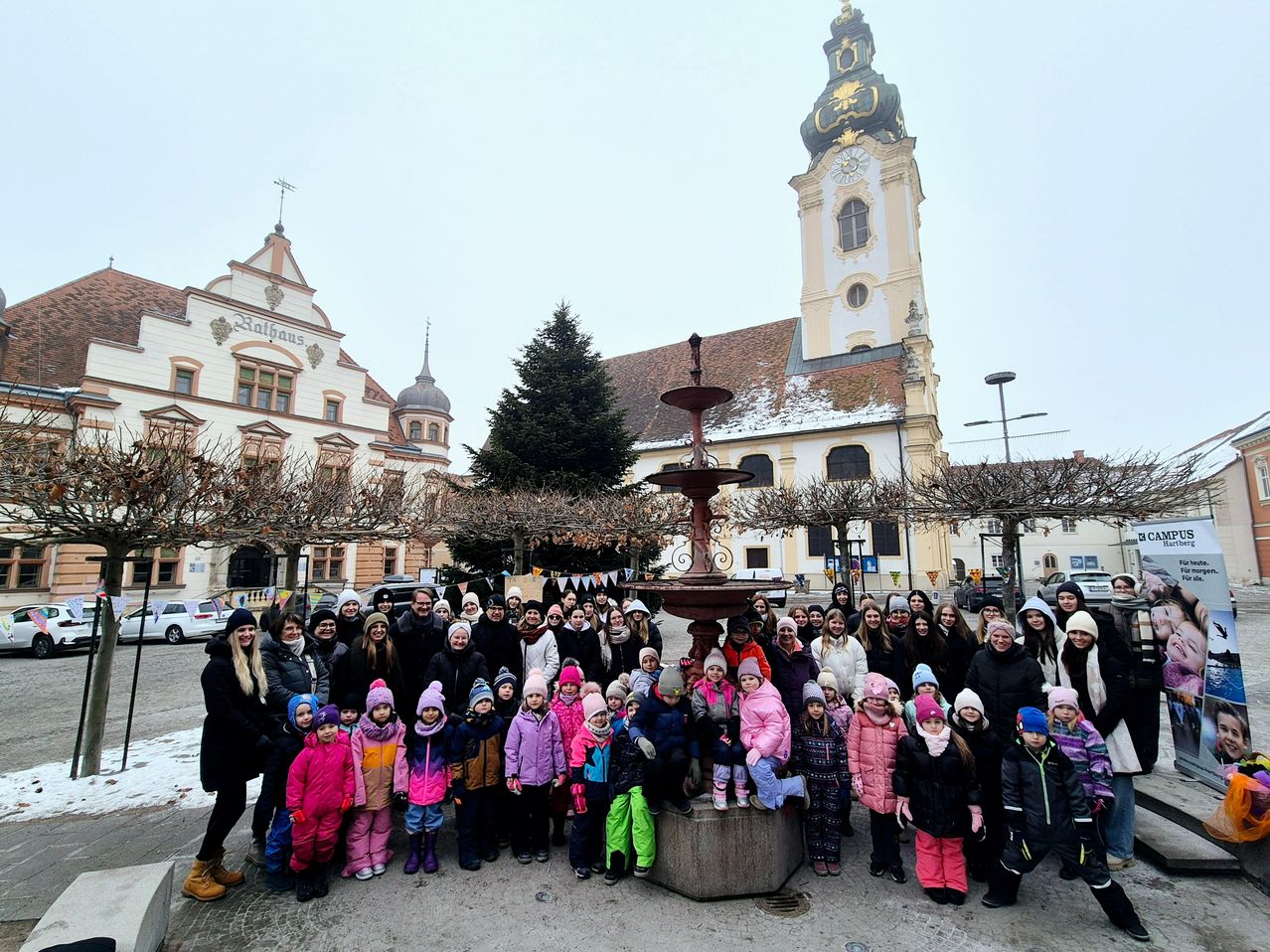 Eine Gruppe von Menschen, einschließlich Kindern, steht vor einem Brunnen und einer Kirche. Sie sind in Winterkleidung gekleidet. Bäume und ein Glockenturm sind im Hintergrund.