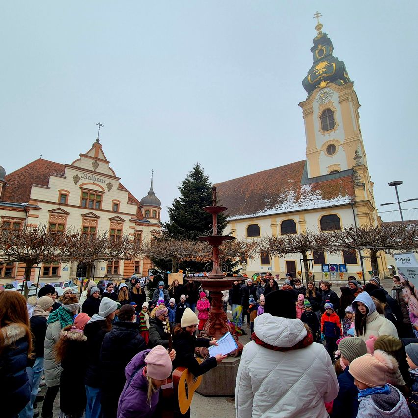 Eine Menschenmenge versammelt sich um einen Brunnen vor einer Kirche. Eine Person spielt Gitarre und liest von einem Blatt Papier. Die Kirche hat einen Turm mit einer Uhr und einer Wetterfahne.