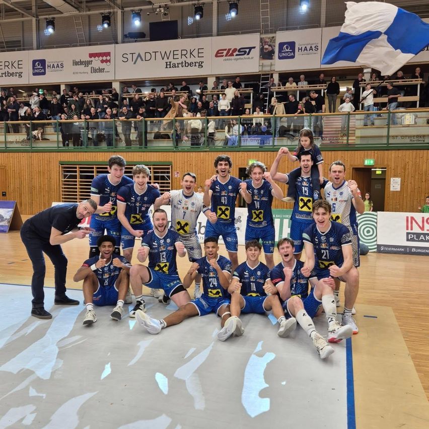 Eine Sportmannschaft posiert für ein Foto auf einem Spielfeld, mit einem Publikum im Hintergrund. Die Spieler tragen blaue Uniformen mit Nummern, und einer hat eine Flagge auf seinen Schultern.