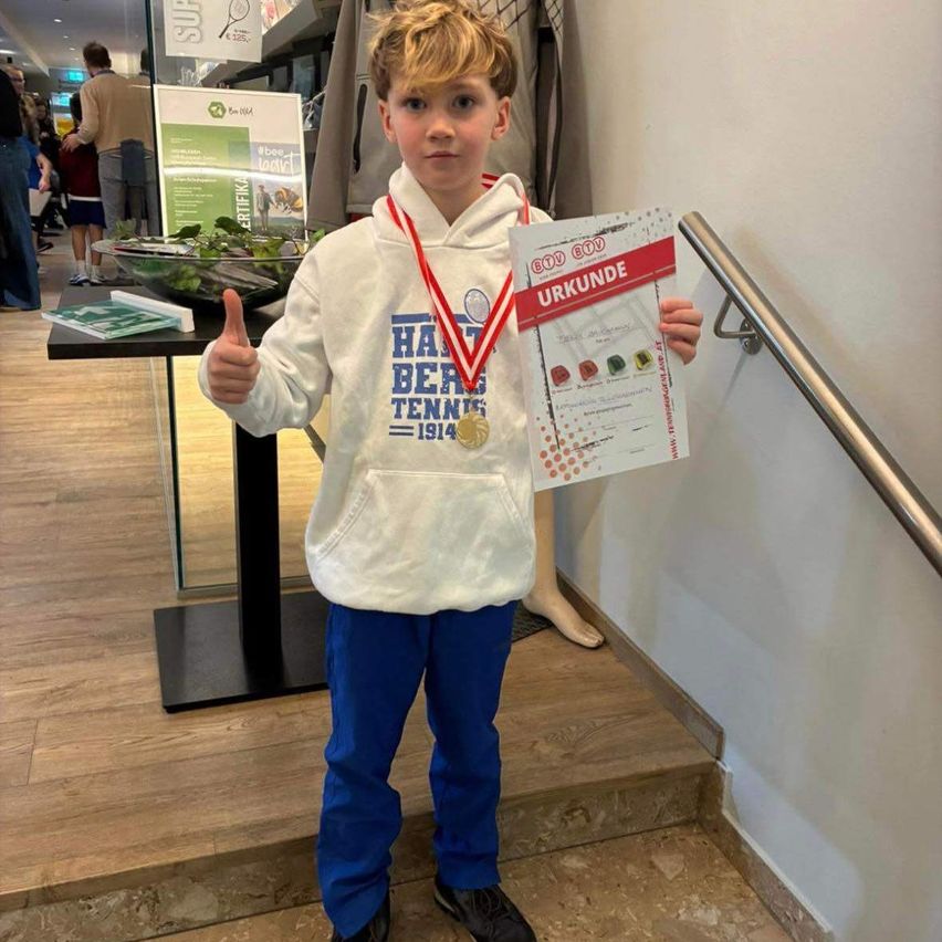 Ein junger Junge mit einer weißen Kapuzenjacke hält ein Zertifikat und zwei Medaillen in der Hand und gibt ein Daumenhoch.