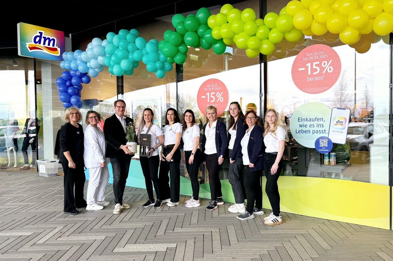 Eine Gruppe von Menschen steht vor einem Geschäft mit Luftballons und Schildern, die einen 15% Rabatt anbieten. Sie halten einen Pokal und lächeln für ein Foto.