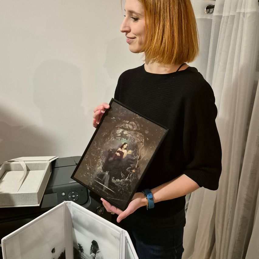 Eine Frau mit roten Haaren hält ein gerahmtes Foto mit der linken Hand und ein offenes Fotoalbum in der rechten. Sie lächelt und scheint für ein Bild zu posieren.