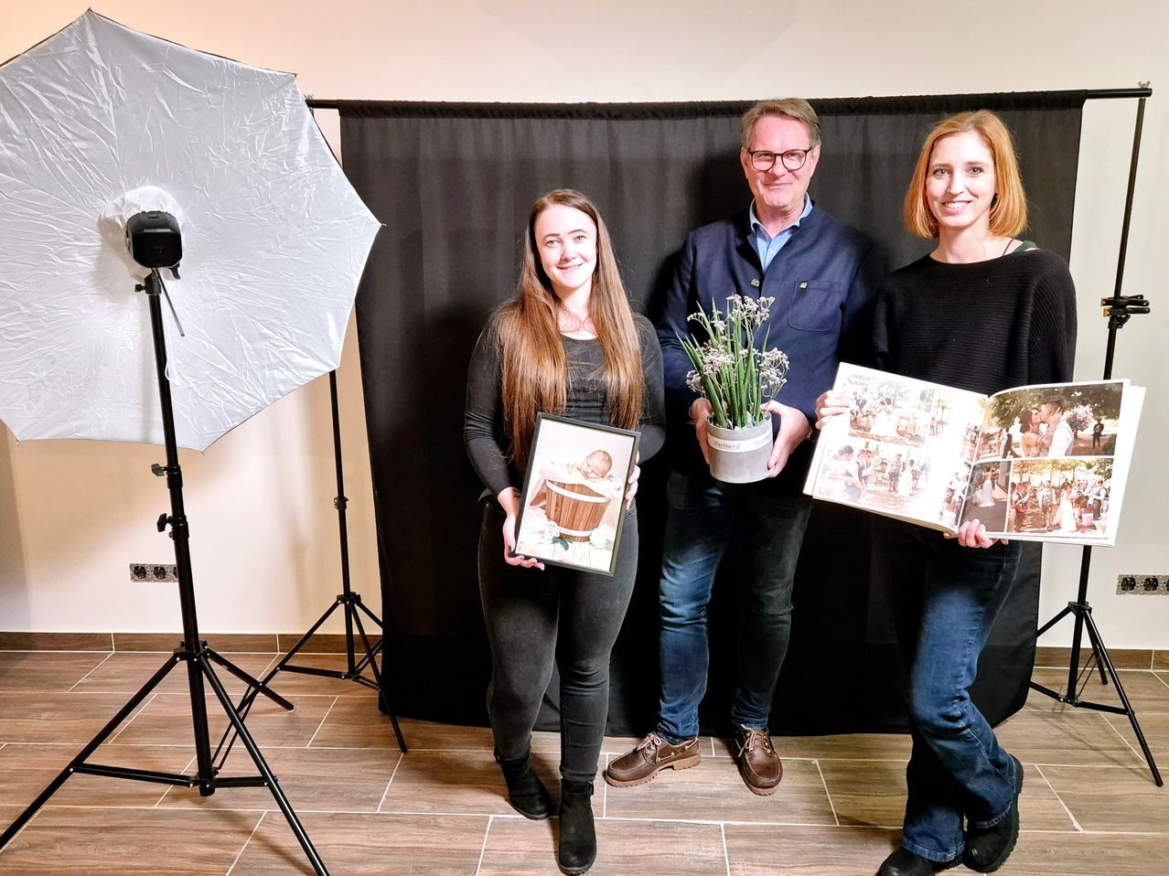 Drei Menschen stehen lächelnd vor einem schwarzen Hintergrund. Eine Frau hält ein Fotoalbum, eine andere ein Babyfoto und ein Mann hält eine Topfpflanze. Dahinter steht ein weißer Regenschirm und ein Stativ.