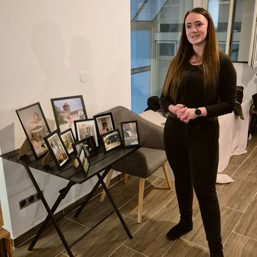 Eine Frau in einem schwarzen Outfit steht neben einem Tisch mit Fotorahmen. Der Raum hat weiße Wände und einen Holzboden.