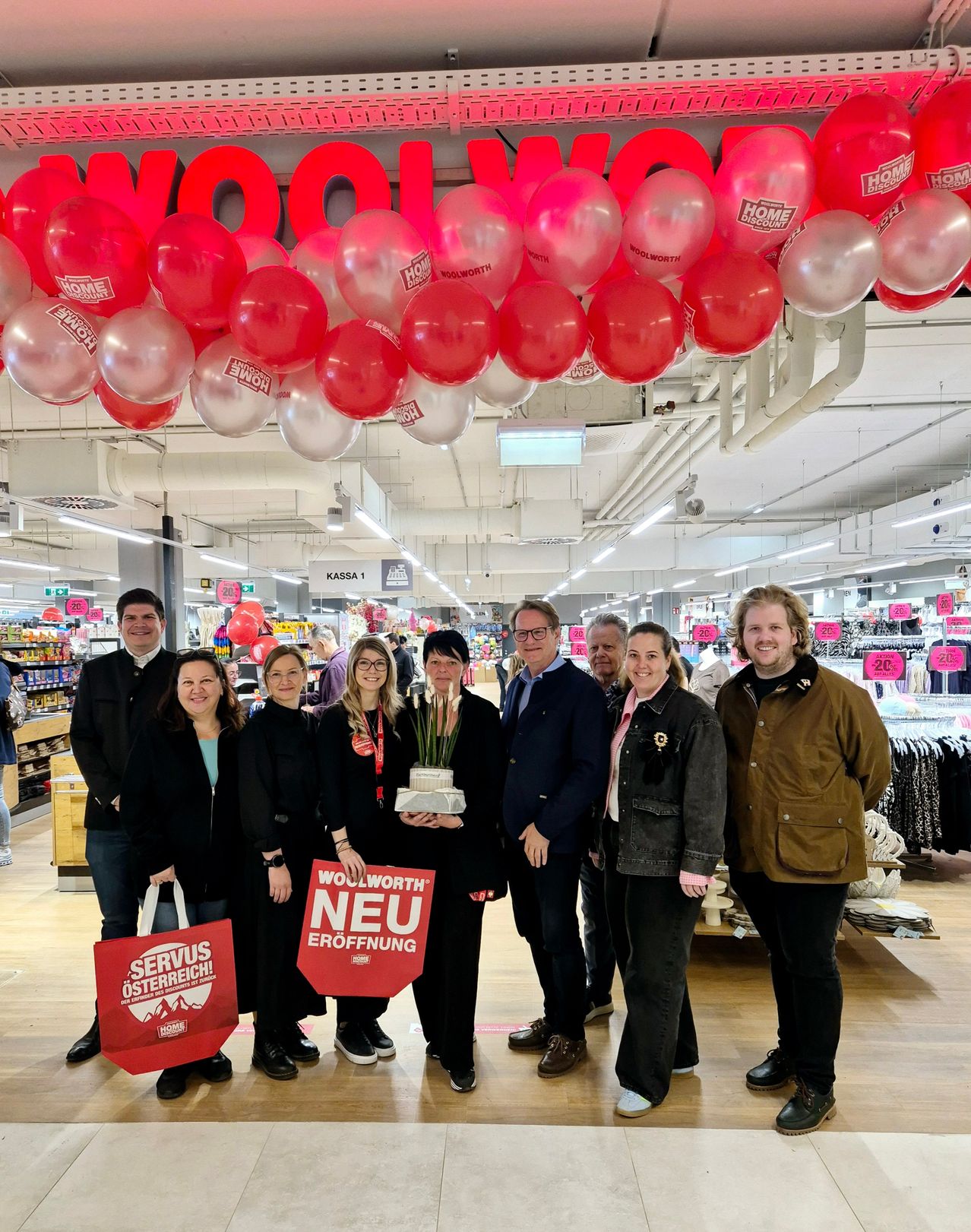 Eine Gruppe von Menschen steht in einem Woolworth-Geschäft, hält Schilder und posiert für ein Foto. Im Hintergrund sind Luftballons und Schilder mit den Worten 'Woolworth' und 'Neue Eröffnung' zu sehen.