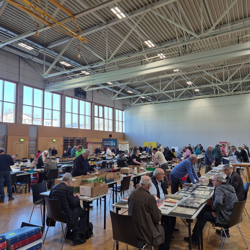 Ein großer Innenraum mit vielen Tischen und Stühlen, an denen Menschen sitzen und stehen und mit Büchern und Kisten arbeiten. Der Raum hat große Fenster und eine hohe Decke mit Beleuchtung.