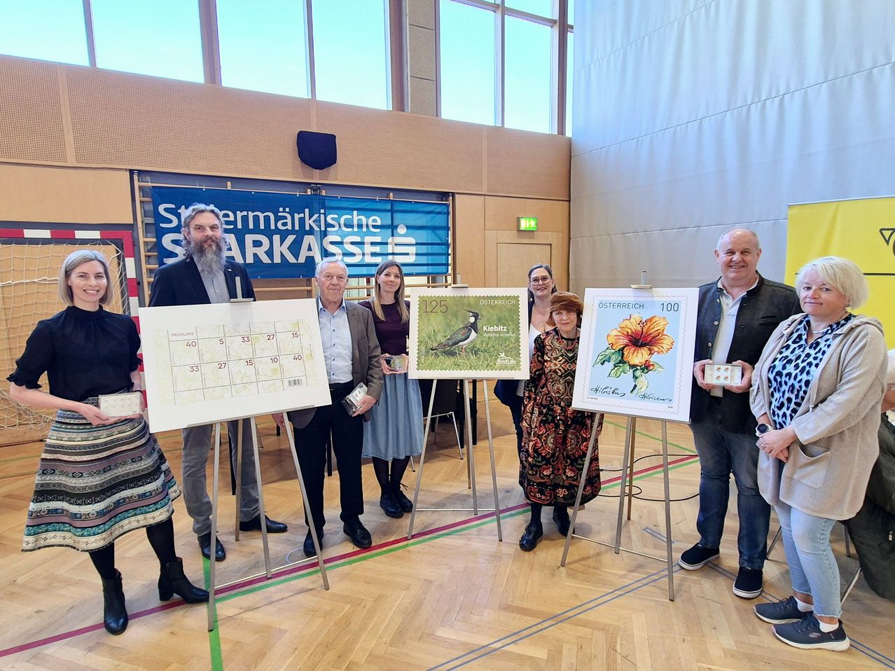 Eine Gruppe von Menschen steht in einem Raum mit Holzböden und zeigt zwei Poster mit Briefmarken. Ein Poster zeigt einen Vogel und das andere eine Blume.