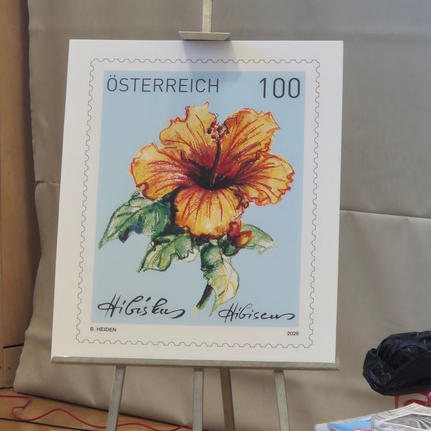 Eine gerahmte Briefmarkenanzeige zeigt eine orange Hibiskusblume mit grünen Blättern. Die Briefmarke stammt aus Österreich und trägt den Namen des Künstlers, B. Heiden, unten.