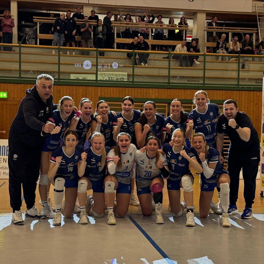 Eine Frauen-Volleyballmannschaft feiert einen Sieg in einer Sporthalle mit einer Menge Zuschauer im Hintergrund.