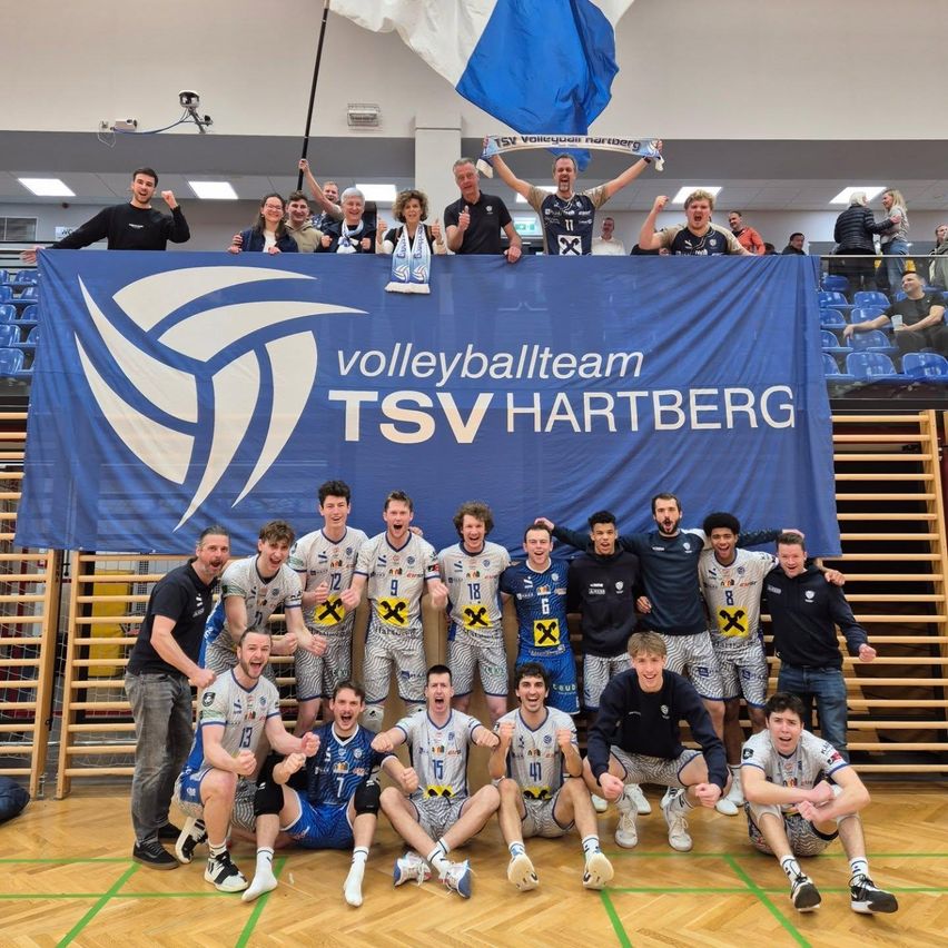 Das Volleyballteam TSV Hartberg feiert einen Sieg, während die Spieler vor einem blauen Banner für ein Foto posieren. Einige Leute stehen hinter dem Banner auf der Galerie.