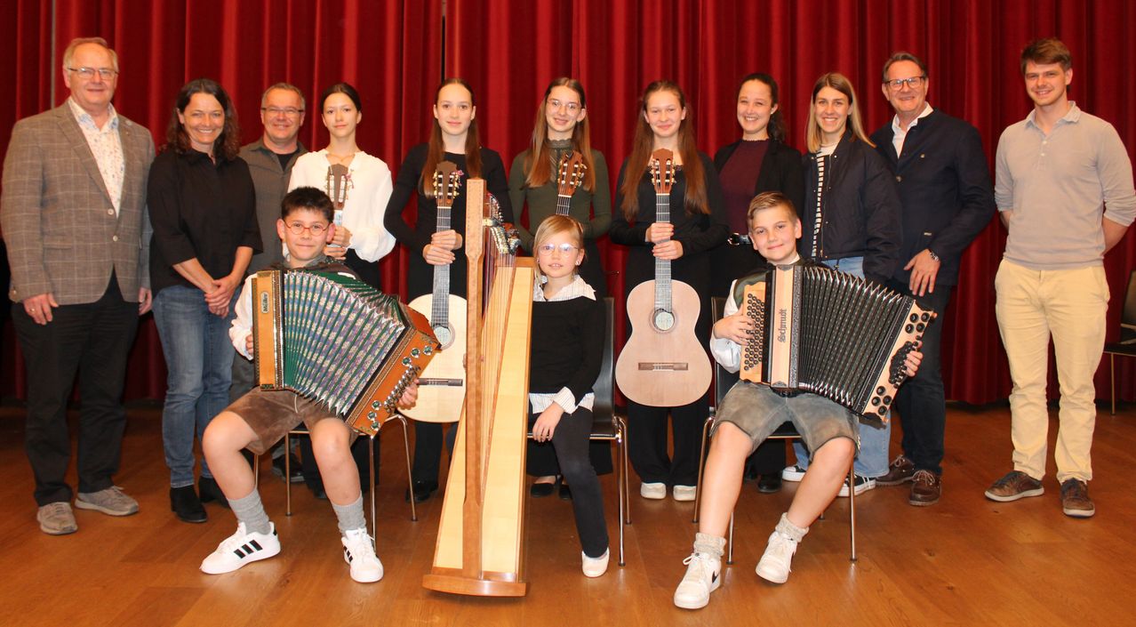 Eine Gruppe junger Musiker, bestehend aus Jungen und Mädchen, posiert mit verschiedenen Musikinstrumenten wie Gitarren, Harfen und Akkordeons vor einem roten Vorhang.