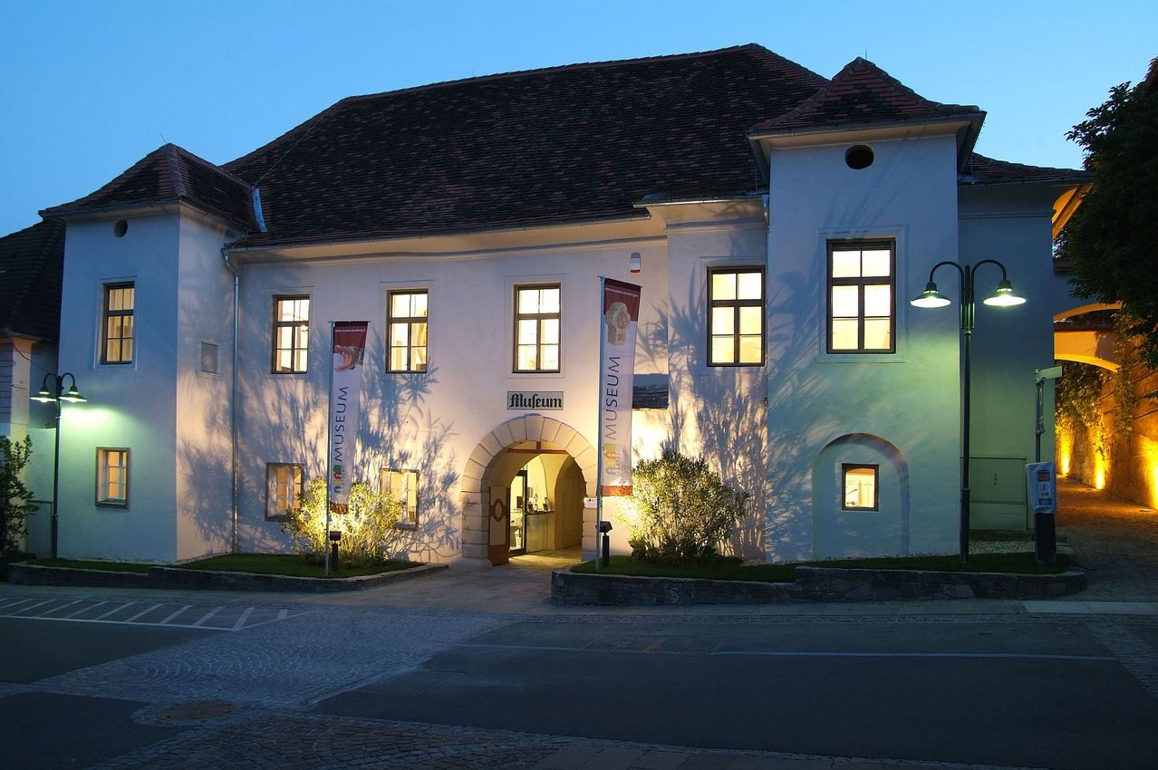 Ein gut beleuchtetes Museumsgebäude mit gewölbtem Eingang, Fahnen und beleuchteten Fenstern in der Dämmerung.