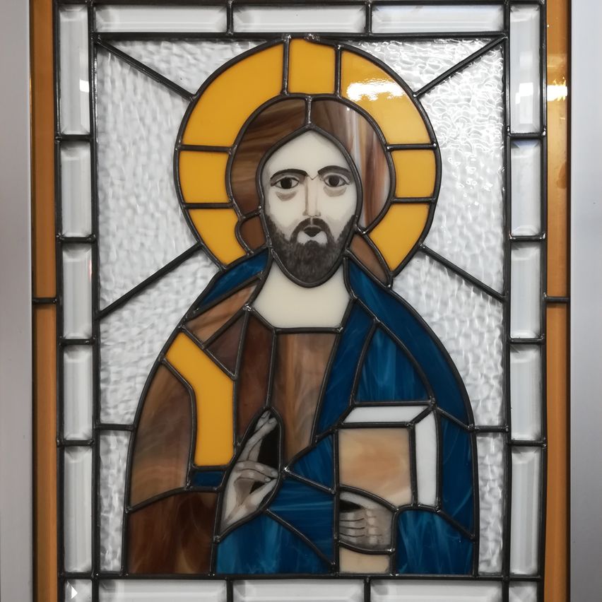 Ein Buntglasfenster zeigt Jesus mit einem Buch in der Hand, umgeben von einem gelben Heiligenschein und einem blauen Gewand.