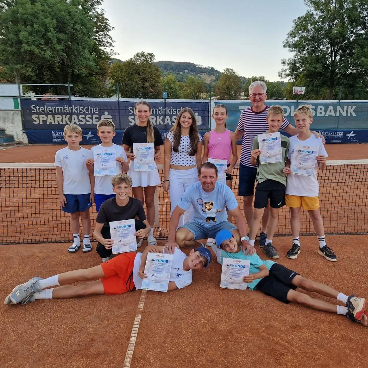 A group of young tennis players and a coach pose for a photo on a tennis court with certificates in hand.