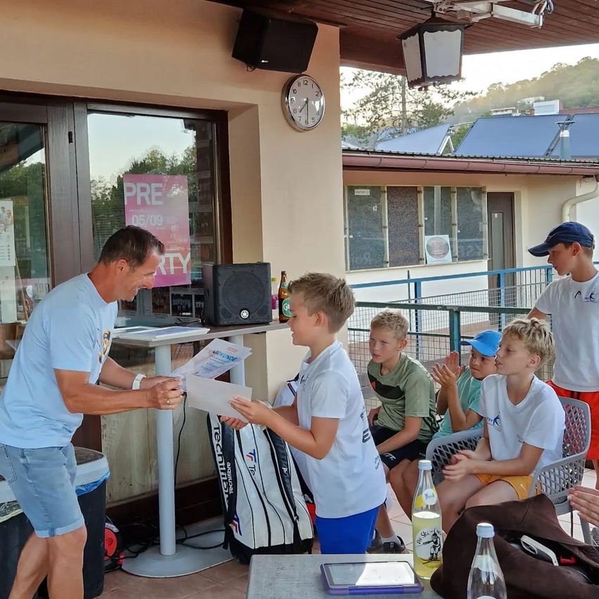 A man hands a paper to a boy in a white shirt, while other boys watch. They are outside near a table with tennis bags. A clock and a sign are visible in the background.