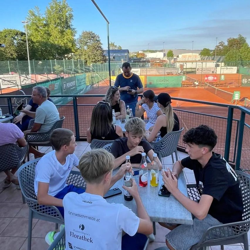 A group of people are sitting around a table on a terrace, possibly at a tennis court. They are engaged in conversation, with some of them holding bottles. The setting is outdoors, with a tennis court visible in the background.