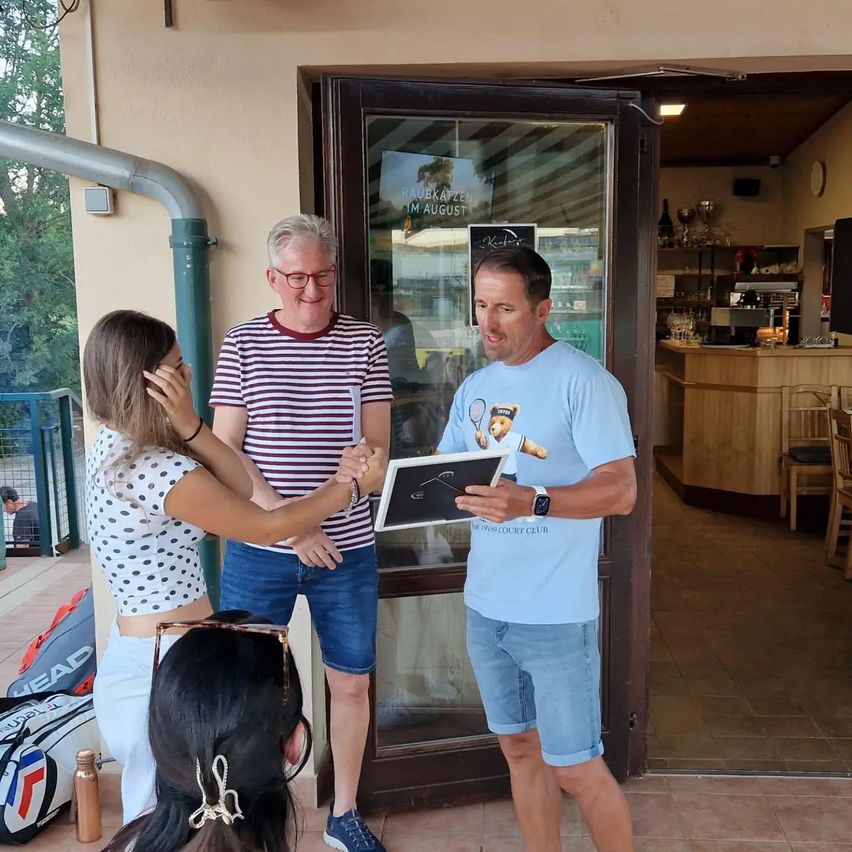 Two men and a woman stand in front of a glass door with a sign that reads 'RAUBZEIT IM AUGUST'. The man on the right holds a framed certificate, and the woman on the left holds his hand. A tennis racket is visible on his shirt.
