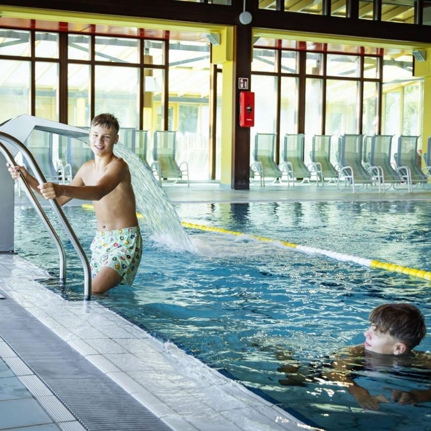 Two boys are swimming in an indoor pool. One boy is climbing out of the pool using the handrail.