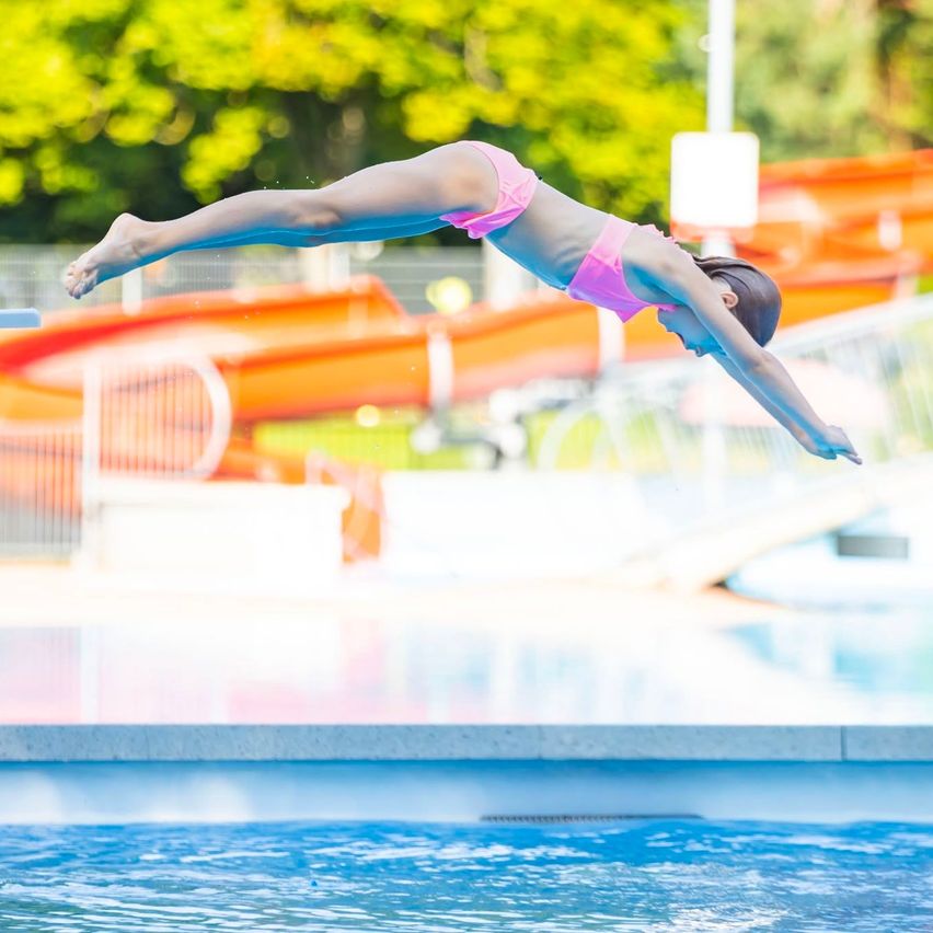 A young girl dives into a swimming pool with a water slide in the background.