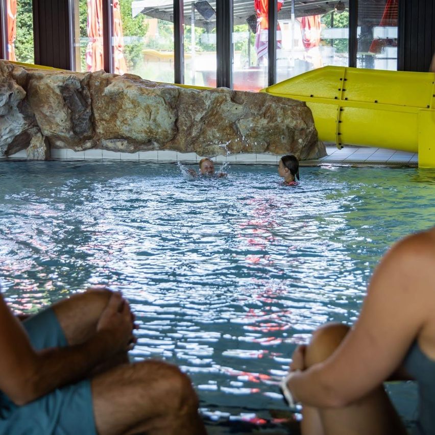 Two children are swimming in a pool with a yellow slide in the background. Two adults are sitting on the side of the pool.