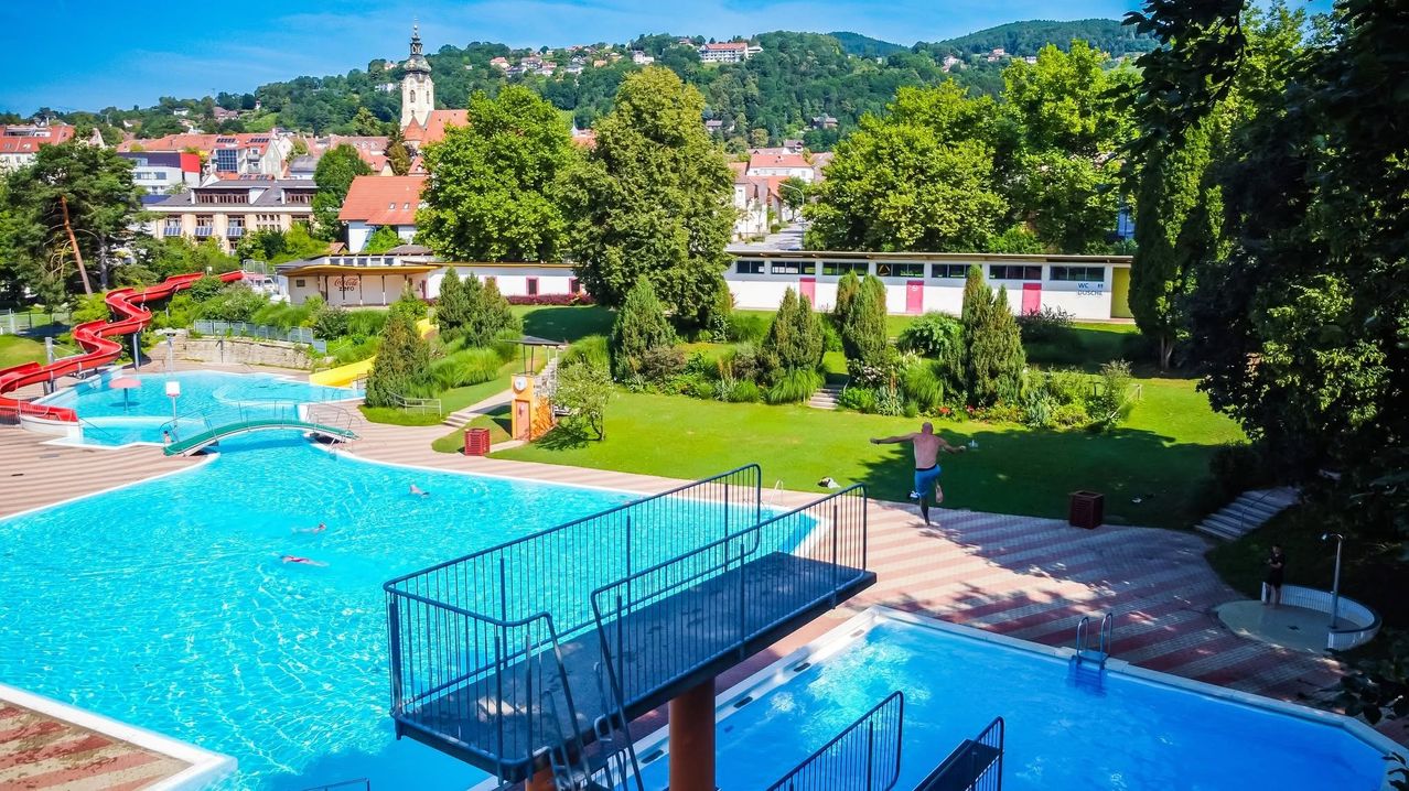 Aerial view of a swimming pool with a man jumping into it, surrounded by lush green trees and buildings in the distance.