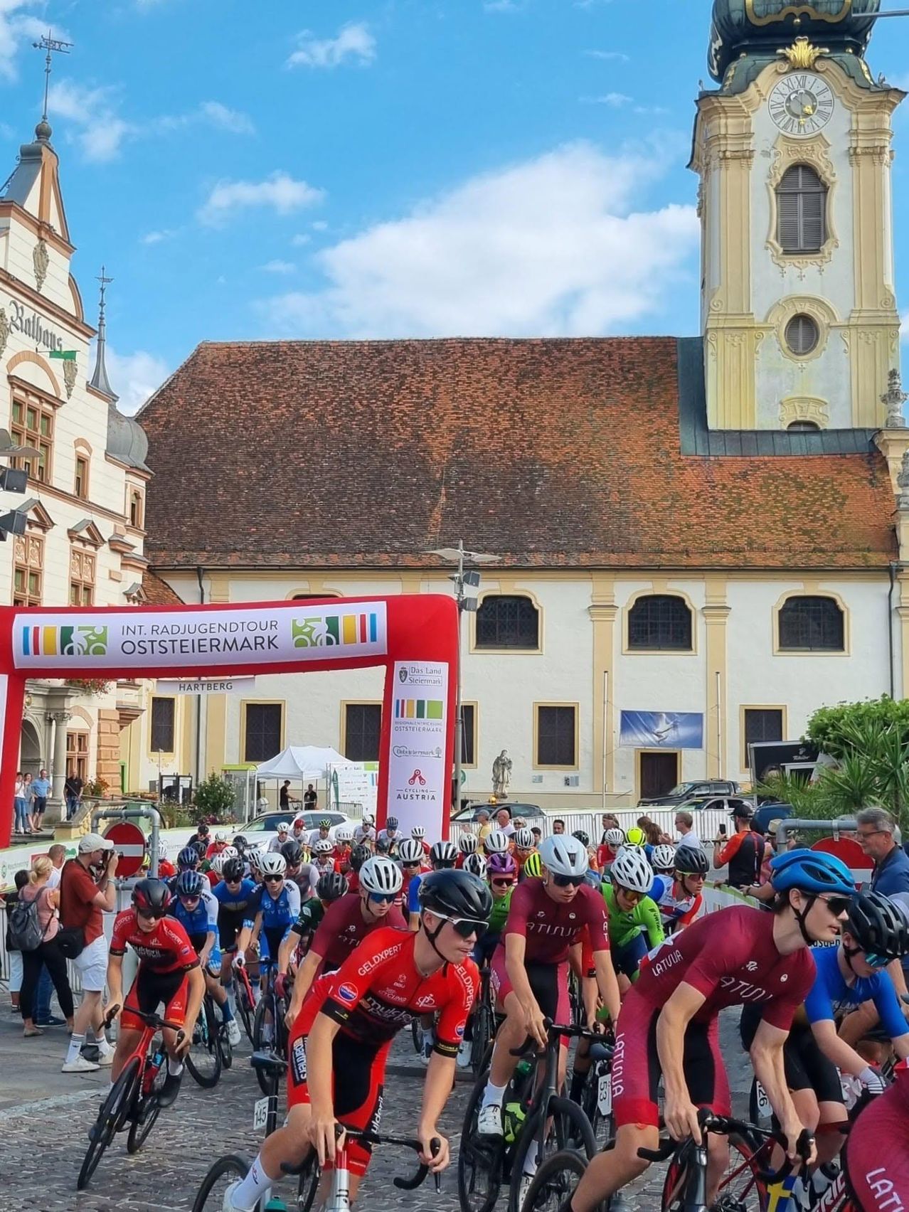 A group of cyclists wearing helmets and cycling jerseys are lined up at the start of a race in front of a historic building.