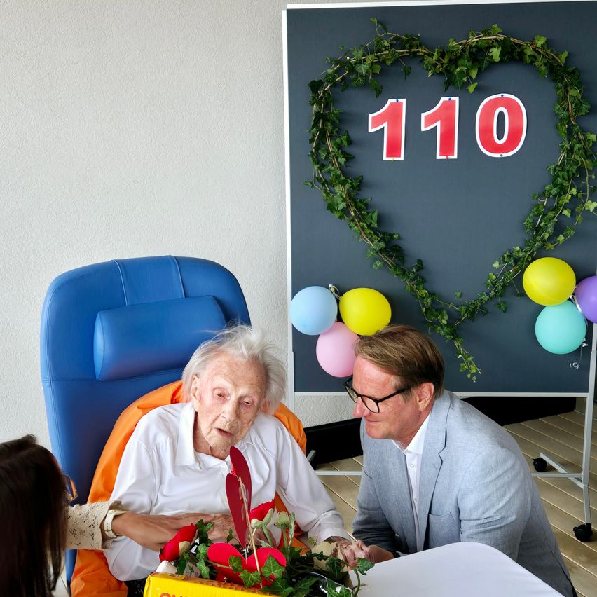An elderly woman in a blue chair receives a bouquet of flowers from a man in a suit. Behind them, a heart-shaped decoration with the number 110 and balloons adorns the wall.