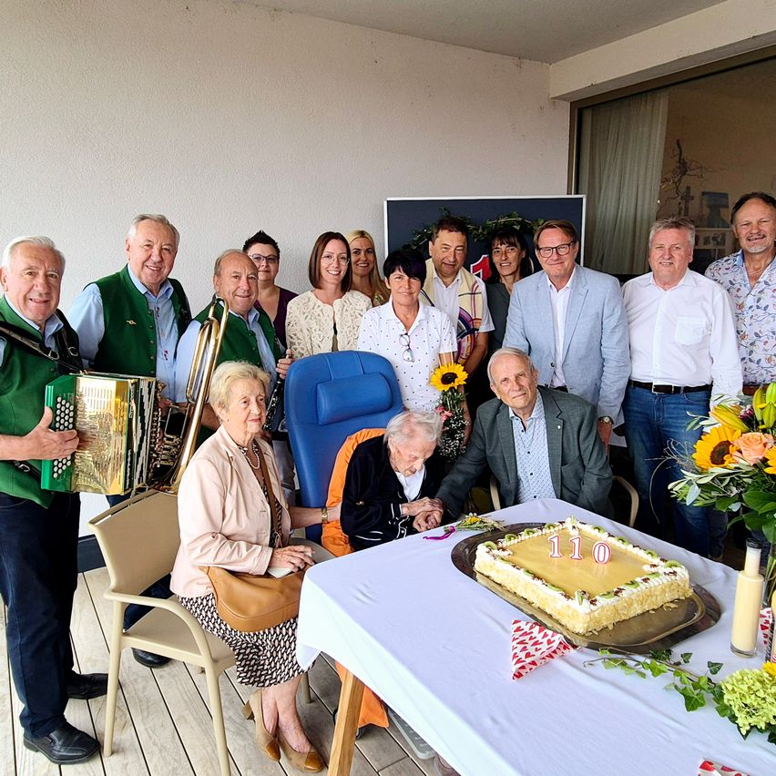 A group of people celebrating a birthday with a cake, flowers, and musical instruments in a room.