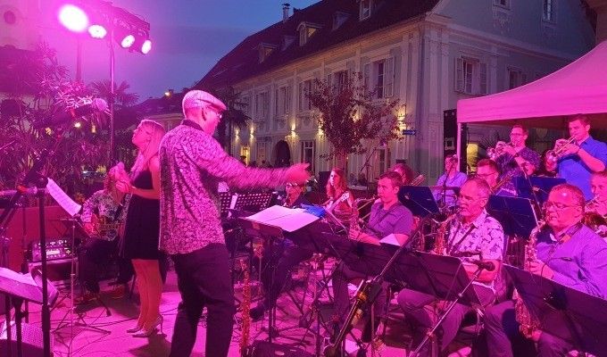 Eine Jazzband spielt bei Nacht auf einer Bühne, beleuchtet von lila Lichtern, mit einem Gebäude und einem Zelt im Hintergrund.
