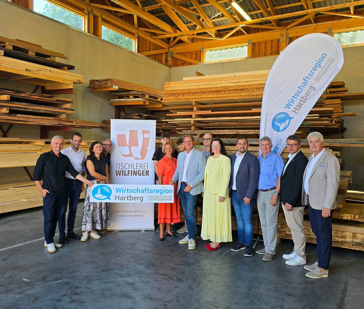 A group of people stand in front of a wooden storage area, holding a banner that reads 'Wirtschaftsregion Hartberg'. They are dressed in business attire, and the setting suggests a formal event or meeting.