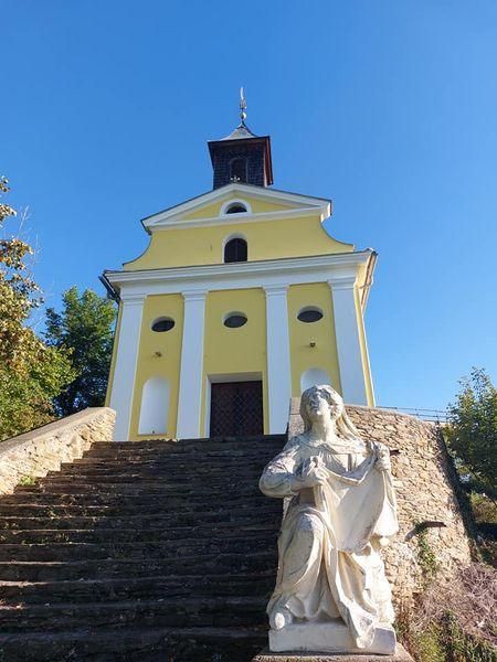 A yellow church with a tower and a statue of a woman in front of it. The church is surrounded by a stone wall and has a staircase leading up to it.